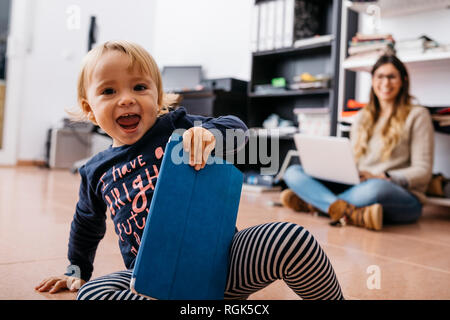 Feliz niña sentada en el suelo en casa con tablet y madre en segundo plano.