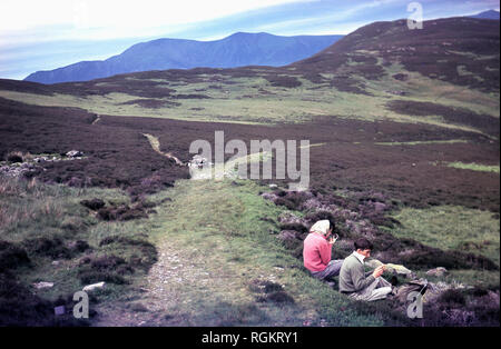 1960, históricos, pareja apoyada en el suelo por un camino mientras caminando por encima de Rydal water, Lake District, Cumbria, Inglaterra, Reino Unido. La virgen, cubierta con un velo, está sentado aplico maquillaje utilizando un espejo, Foto de stock
