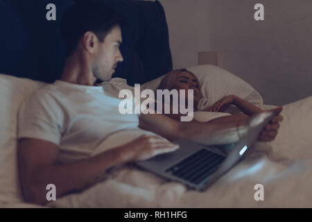 Joven sospechoso con ordenador portátil y mirando novia dormir en la cama en la noche Foto de stock