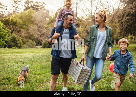 Familia feliz con el perro caminando hacia el picnic en el jardín. El hombre lleva a su hija sobre los hombros con hijo mujer sosteniendo la mano y llevando un