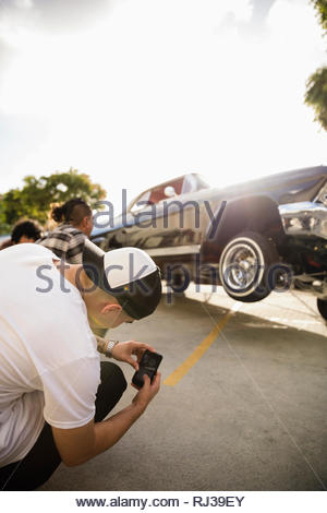 Latinx joven con cámara teléfono fotografiar bajo rider rebotando en coche aparcamiento