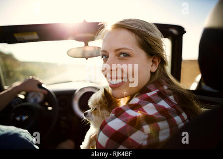 Retrato de una sonriente joven mujer sentada en el asiento del pasajero de un automóvil convertible con su pequeño perro.