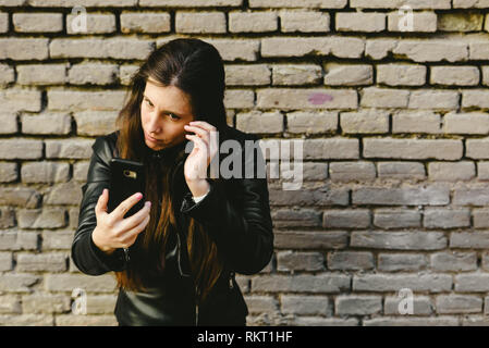 Chica joven con ayuda de la pantalla de su teléfono móvil como un espejo de maquillaje para poner y quitar la piel de su rostro, y ella obtiene bastante para una fecha con ella Foto de stock