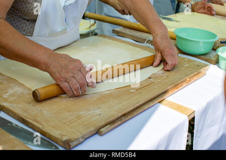 Viejo arrugado manos de anciana rodando la masa con rodillo sobre una tabla para cortar madera. Concepto: beneficios de cocinar en casa, vida activa i Foto de stock