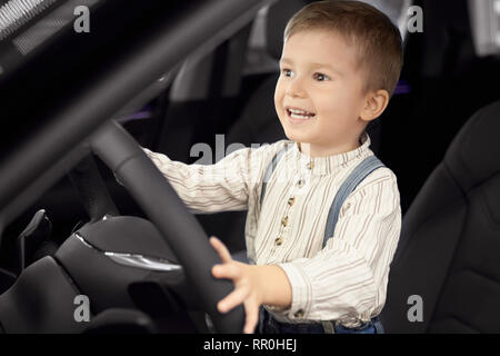 Lindo, guapo chico sentado en el asiento del conductor de coche camarote. Niño feliz tomados de las manos en el volante, apartar la mirada, sonriendo. Concepto de concesionario de coches showroom. Foto de stock