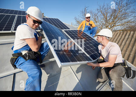 Los trabajadores varones instalación fotovoltaica solar sistema. Electricistas azul elevación módulo solar en el techo de la casa moderna. Concepto ecológico de energía alternativa.