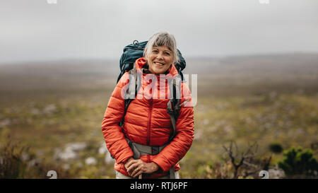 Retrato de una mujer sonriente caminante de pie en la cima de una colina en un día de invierno. Senior mujer vistiendo chaqueta y mochila trekking sobre una colina.