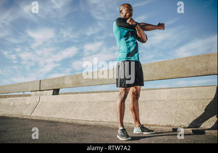Hombre afroamericano en ropa deportiva estirando sus brazos. Atleta Masculino de calentamiento antes de correr por la mañana en la ciudad. Foto de stock