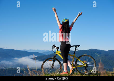 Vista posterior del deportivo ciclista femenina de pie con sus manos con bicicleta amarilla en la cima de la montaña, con casco, disfrutando de neblina matinal en el valle. La actividad al aire libre, el concepto de estilo de vida. Espacio de copia