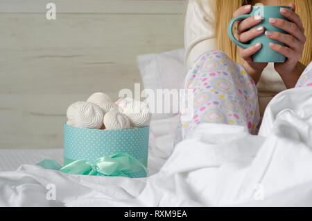 Mujeres manos sosteniendo una taza con bebida en la cama. Melcochas y merengues en una caja. Buena mañana y concepto de relajación. Espacio de copia