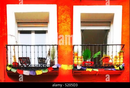 Coloridos balcones de dos ventanas adyacentes en hierro forjado, ornamentado con decoración multicolor bunting contra una fachada de color rojo-naranja brillante. Dos pequeñas sillas de madera en un balcón y macetas con plantas Foto de stock