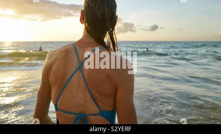 Mujer con el cabello mojado vestido de azul bañador apartar la mirada de la cámara hacia la puesta del sol sobre el Océano Pacífico