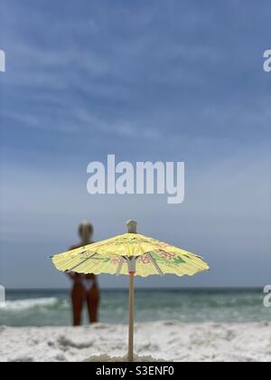 Selecciona el enfoque en un paraguas en miniatura con una mujer y una playa borrosas antecedentes Foto de stock
