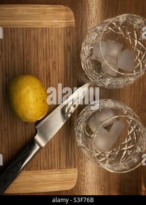 Hacer ginebra y tónico - dos vasos cortados desde arriba con cubitos de hielo junto a un cuchillo y limón en una tabla de cortar Foto de stock