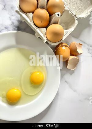 Huevos crudos. Aislados. Huevos agrietados en un plato blanco. Caja de huevos con huevos enteros en el fondo. Imagen de stock. Foto de stock