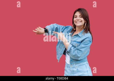 Retrato de feliz hermosa mujer joven morena en jeans con maquillaje permanente de estilo informal, mostrando y señalando a su Smart Watch con gran sonrisa. Foto de stock