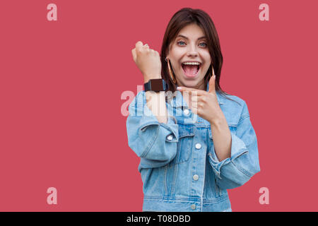 Retrato de sorpresa feliz hermosa mujer joven morena en jeans estilo casual mostrando permanente y apuntando a la pantalla de su reloj inteligente con shock Foto de stock