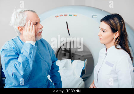 Paciente Senior queja de dolor de cabeza a su médico mientras está sentado en la mesa del escáner. Foto de stock