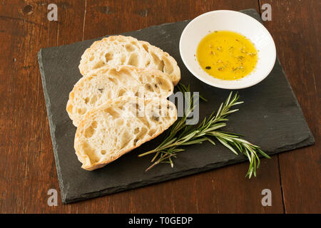 Tres rebanadas de pan ciabatta sobre una placa de pizarra con resortes de romero y un tazón de hierba-infundido el aceite de oliva virgen extra en una mesa de madera angustiado.