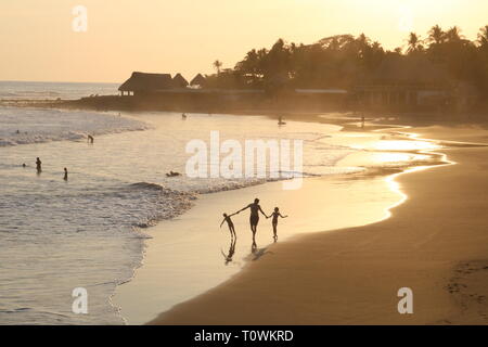 Silueta de un padre y sus dos hijos jóvenes caminando en una playa tropical al atardecer.