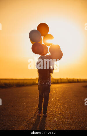 Niña caminando con globos de colores en la carretera en el campo al atardecer. Libertad de verano y viajes. Concepto de esperanza. Emociones y sentimientos. Foto de stock