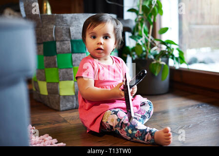 Retrato de lindo bebé niña sentada en el suelo en casa manteniendo el control remoto