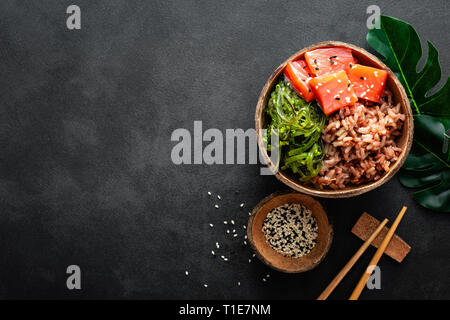 Poke bowl con salmón, pescado crudo chuka ensalada y arroz en cuencos de coco sobre fondo negro Foto de stock