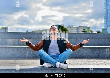 Retrato de joven chico freelance en chaqueta de cuero y zapatillas, relajación y meditación en la calle. El hombre se sienta en las escaleras de una gran ciudad en pos de zen