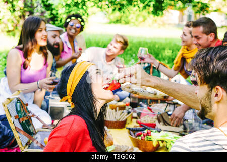 Feliz amigos haciendo un picnic en el jardín al aire libre - moda joven gente divirtiéndose comiendo y bebiendo, mientras están sentados en el césped de la naturaleza Foto de stock
