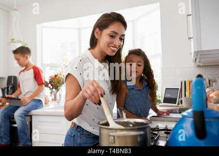 Madre e hija joven preparando la comida en la encimera de la cocina, pre-teen hijo sentado en el fondo