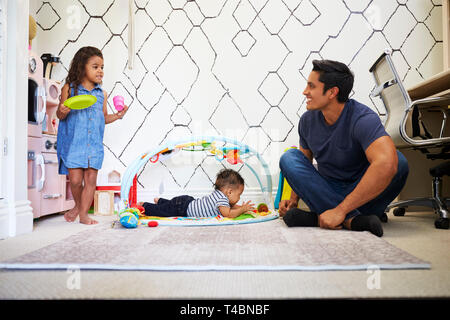Niña jugando tea party con papá, sentados en el suelo, hermano bebé sobre una colchoneta para jugar junto a ellos Foto de stock