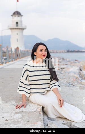 Mujer morena vestida de moda náutica, sentado en las rocas con faro y las  montañas como telón de fondo borroso. Composición vertical Fotografía de  stock - Alamy