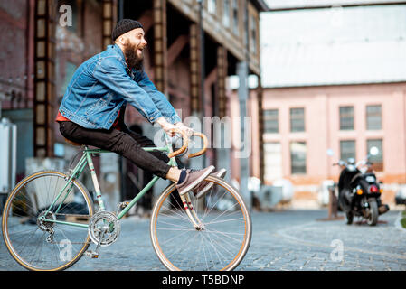 Hombre elegante como un loco hipster divirtiéndose, montando bicicleta retro afuera, en el fondo urbano industrial Foto de stock