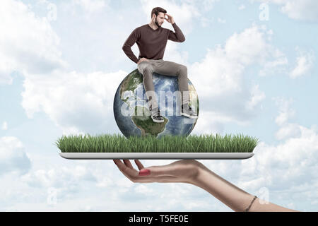 Mano sujetando hembra pequeño hombre en ropa casual sentado sobre el globo terrestre sobre el pasto verde modelo con fondo de cielo azul