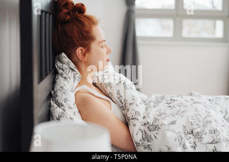 Joven pelirroja mujer relajante sentarse en la cama contra la almohada, con los ojos cerrados y la luz del sol que entra a través de la ventana detrás