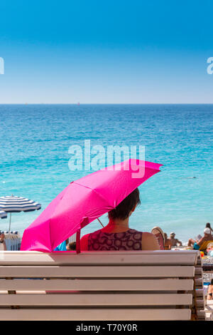 Mujer sentada en un banco con paraguas rosa mira al mar en Niza, Sur de Francia. Foto de stock