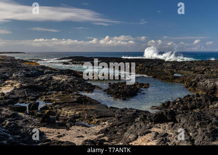 Del arricife en playa volcánica cerca de Kona, en la isla más grande de Hawai. Rocas Negras en primer plano, con olas de marea entrante. Océano Azul cielo y nubes en