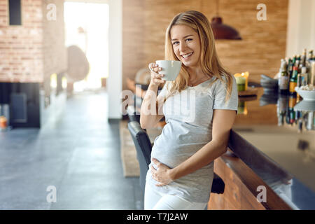 Feliz sonriente joven embarazada relajante apoyado contra una barra de bar con una taza de té caliente acunando a su bebé golpee en su mano como ella bonos con h