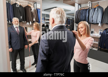 Anciano hombre tratando de traje formal en la tienda de ropa femenina y adaptar la medición de la longitud de la manga. Elegante caballero vistiendo elegante traje inteligente mirando en el espejo y elección. Concepto de montaje.