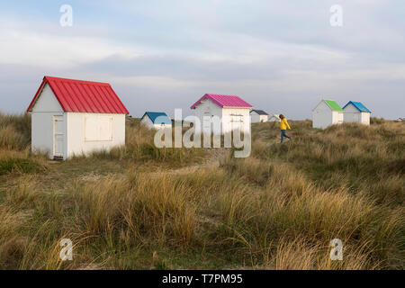 Mujer de chaqueta amarilla caminando sobre hierba en hilera de cabañas de playa de madera blanca con techos coloridos en Gouville-sur-Mer, Normandía, Francia