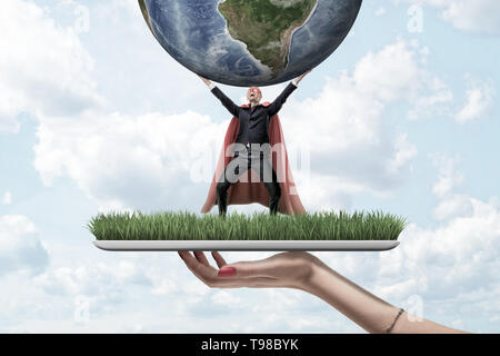 Mano femenina sosteniendo con placa de hierba pequeño empresario vestidos de rojo y el manto de máscara de superman sosteniendo globo terrestre sobre fondo de cielo azul