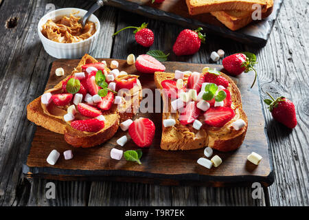 Sándwiches de pan tostado con mantequilla de cacahuete, rodajas de fresas y marshmallow sobre una tabla de cortar de madera rústica en una vieja tabla, vista horizontal