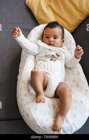 Directamente encima de la vista de feliz emocionado African Baby Boy en lindo capullo bodysuit acostado en seguridad en el sofá y agitando brazos pequeños Foto de stock