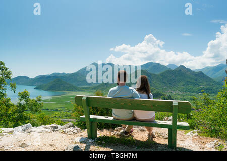 La pareja se sienta en un banco y admira las vistas al lago y a las colinas en un día soleado Foto de stock