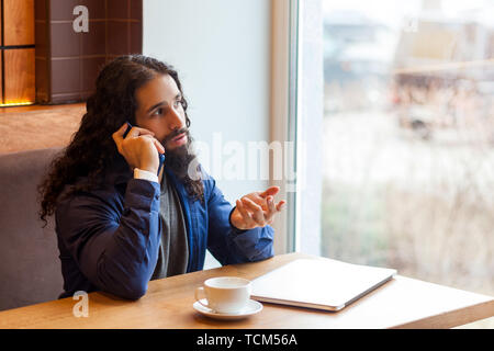 Retrato de graves inteligencia guapo joven adulto hombre freelancer en estilo casual sentado en la cafetería con un portátil, hablando por teléfono, mirando a otro lado, Foto de stock