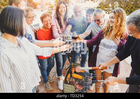 Familia Feliz aclamaciones y brindando con vino tinto en vidrio parrillada - Personas con diferentes edades divertirse beber y cocinar carne Foto de stock