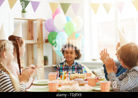 Retrato de afroamericanos sonriente niño usando crown sentado en la tabla mientras celebrando un cumpleaños con amigos, espacio de copia