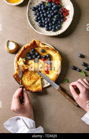 Joven manos comienzan cortando bebé holandés pancake en placa de cerámica servidos con Blackberry y grosellas bayas, tazón de miel, jarra de crema, vintage Foto de stock