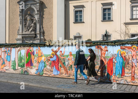 Pascua de Viena, la vista de una joven pareja caminando por una colorida ilustración de la Pasión de Cristo junto a la Schottenkirche en el centro de Viena. Foto de stock