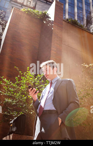 Ángulo de visión baja del empresario utilizando smart phone mientras está de pie contra edificios en la ciudad Foto de stock